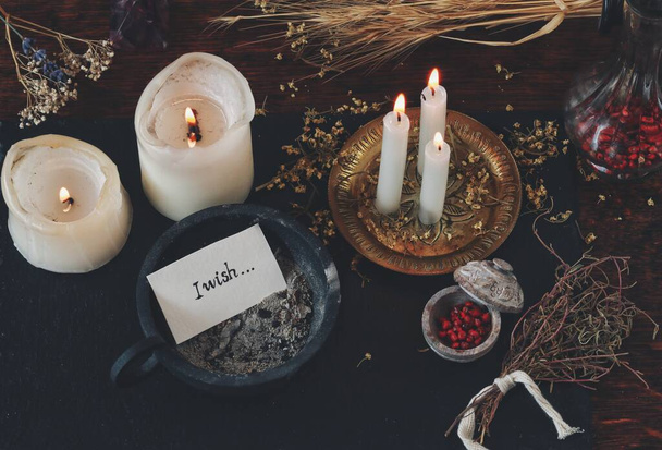 Peace of paper with words I wish written on it, put in cast iron candle holder with ashes in it, on wiccan witch altar. Casting a spell for wish to come true, manifesting. Burning candles, dried herbs in the background of dark wooden table. - Photo, Image
