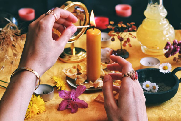 Bruja bruja wicca (con joyas vintage) encendiendo una vela amarilla en su altar de celebración del Solsticio de Sabbat a mediados del verano de Litha llena de flores y cosas al azar correspondientes al Sol. Sostiene una cerilla en sus manos para encender una vela
 - Foto, imagen