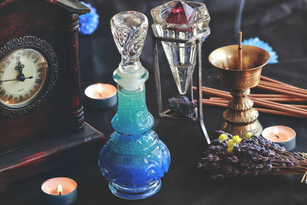 Lucid dreaming good night potion on a witch wiccan altar. Blue colored liquid in a beautiful vintage potion bottle. Slightly blurred background with burning candles, incense, crystals, lavender, clock - Photo, Image