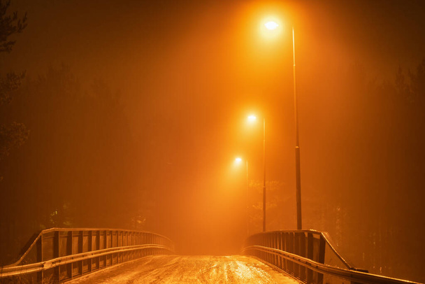 Foggy view of small transport bridge with lighting three streetlights at the right side. Blurry autumn mist colored in orange by lights air hides details. Umea city, Vasterbotten, Northern Sweden - Photo, Image