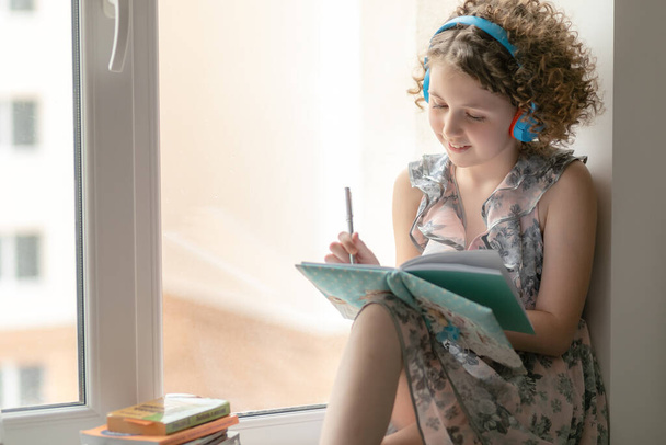 Curly girl in headphones sits at the window. Music helps to combine study and relaxation. A teenager smiles and makes notes in a notebook. Isolation is no reason to be sad - Фото, изображение