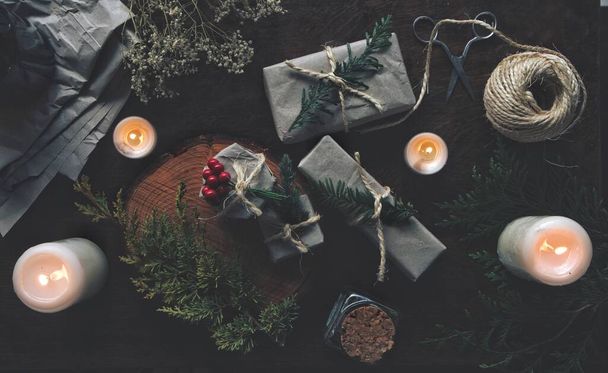 Karácsonyi téli napforduló (Karácsony) témájú lapos feküdt ajándékok csomagolva szürke papír szüret csomagolás fonallal. Szárított baba leheletvirág, örökzöld faágak, égő fehér gyertyák sötét fa asztalon. A gyártási folyamat közepén - Fotó, kép