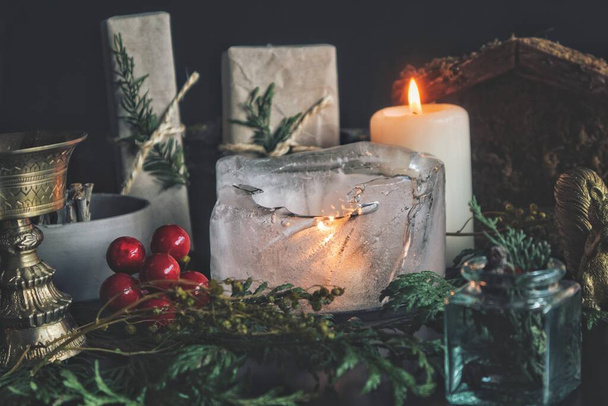 Close up of a candle burning in the ice. Festive Yule winter solstice (Christmas) set up on wiccan witch altar, filled with nature items, like evergreens, gifts, presents, white burning candle, gold incense burner, red holly berries - Photo, Image