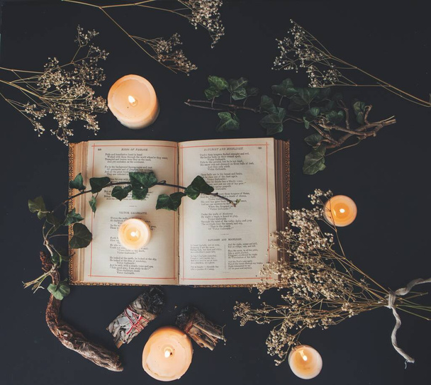 A flat lay of an open vintage poetry book on black table surface with white lit burning candles and dried baby's breath flowers. Dark romantic cozy feel with a branch of ivy on top of the pages and sage smudge sticks near by. Square image photo - Foto, immagini