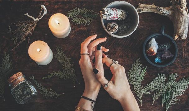 Witches hands on a table ready for spell work. Wiccan witch altar filled with sage evergreen branches herbs crystals and burning white candles. Wearing vintage jewelry on her hands - Photo, Image