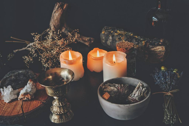 Wiccan witch altar, messy working space with ingredients for a spell. Dried plants nature elements branches and lavender on a table with burning candles, sage smudge sticks and gold brass incense holder - Photo, Image