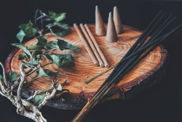 Examples of various types of incense - long black sticks, short hand made sticks and  small triangular cones. Black background, placed on a wooden plate cutting with ivy plant branch laying next to it with slightly blurred background. - Photo, Image