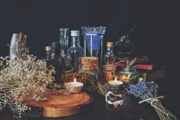 Wiccan witch aptekarz - różne składniki, mikstury i suszone butelki ziół i słoiki do magika, umieszczone na ołtarzu. Ciemne, czarne tło z kwiatami oddechu dziecka, lawenda, palenie zapalonych białych świec, roczniki książek i drewniany kawałek pnia drzewa - Zdjęcie, obraz