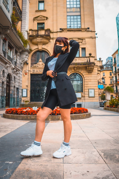 Fashion photo shoot of the new normal, a young brunette entrepreneur with a face mask, coronavirus pandemic. Photoshoot in the city with social distance - 写真・画像