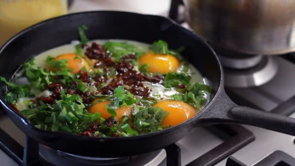 Zbliżenie gotowania przez człowieka jajek smażonych z posiekanym szpinakiem i suszonymi pomidorami w czarnym żeliwnej patelni, selektywne skupienie - Materiał filmowy, wideo