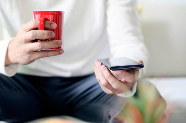 Oříznutý obrázek muže držícího červený šálek, když sedí doma a používá smartphone. - Fotografie, Obrázek