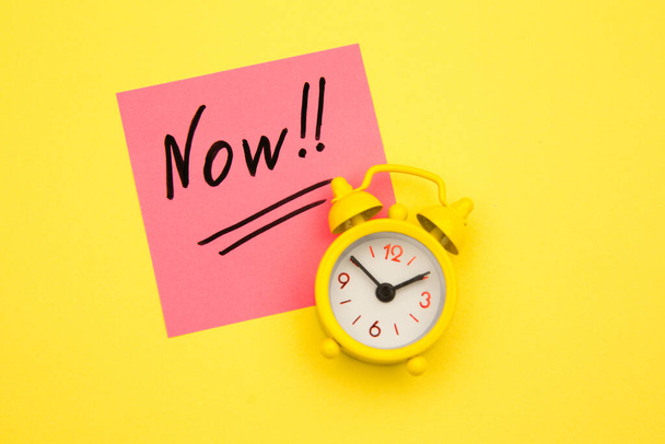 Sticker with handwriting the word NOW on alarm clock on yellow background with copy space using as stop procrastination, self discipline or motivation to complete task - Φωτογραφία, εικόνα