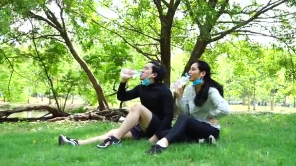 Nastolatki pary noszą stroje sportowe, nosić maski i chronić przed Covi19 Choroby, pić wodę i iść ćwiczyć w parku, Siedząc i odpoczywając, rozmawiać razem. - Materiał filmowy, wideo