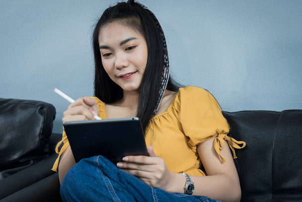 Eine junge Studentin nutzt Tablets und sitzt zu Hause auf der Couch. Eine Teenager-Frau verbringt ihre Zeit während einer Covid-19-Pandemiestudie zu Hause. Online- und Fernstudienkonzept. - Foto, Bild
