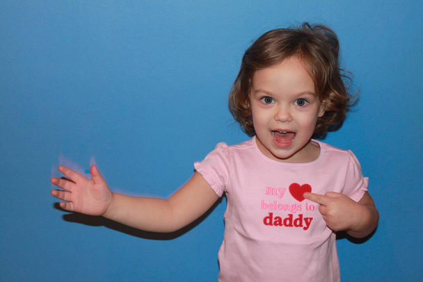 Блондинка в рожевій футболці посміхається, пишучи: "Моє серце належить татусеві" вказує однією рукою на символ серця і хвилі з іншою. Концепция дня отца, день рождения отца, любовь дочери. Блакитний фон - Фото, зображення