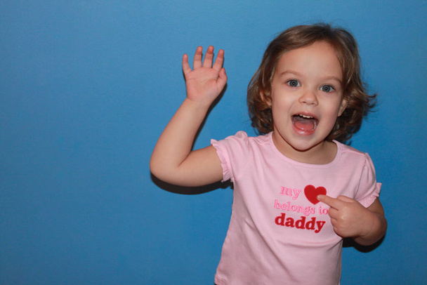 Ragazzina bionda sorridente in t-shirt rosa, scritta: "Il mio cuore appartiene a papà" indica con una mano il simbolo del cuore e saluta con l'altra. Concetto di festa del papà, compleanno del padre, amore della figlia. Fondo blu
 - Foto, immagini