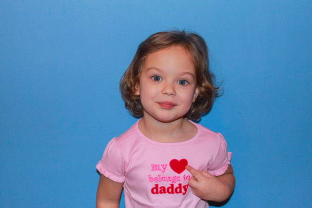 Rubia niña sonriente en una camiseta rosa, con letras: "Mi corazón pertenece a papá" señala el símbolo del corazón. Concepto del Día del Padre, cumpleaños de papá, amor de la hija
 - Foto, imagen