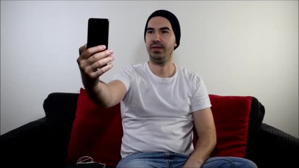 joven macho sentarse en sofá home content creator hold smartphone portátil mano charla selfie sonrisa - Imágenes, Vídeo