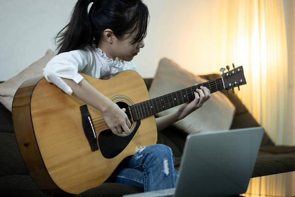 Feliz chica adolescente asiática está practicando tocar la guitarra y estudiar música, cursos de enseñanza de guitarra en línea con ordenador portátil, buscar y aprender en línea, la educación, el desarrollo del conocimiento de Internet
 - Foto, imagen