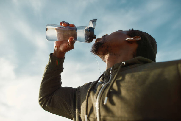 Taking a break. Young african male runner in sportswear drinking fresh water from a bottle while standing against blue sky - Fotoğraf, Görsel