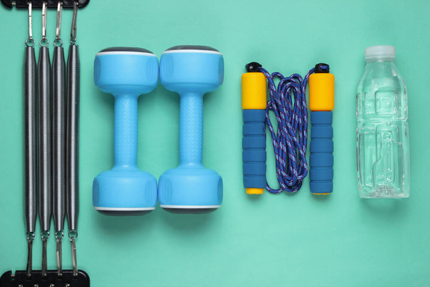 Flaches Laien-Fitness-Konzept. Kurzhanteln, Springseil, Wasserflasche, Expander. Sportgeräte auf blauem Hintergrund. Ansicht von oben - Foto, Bild