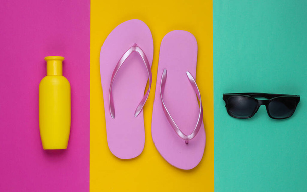 Accessori da spiaggia. Infradito rosa spiaggia alla moda, bottiglia di crema solare, occhiali da sole su sfondo di carta colorato. Piatto. Vista dall'alto - Foto, immagini