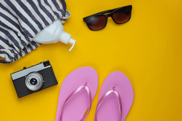 Summer still life. Beach accessories. Fashionable pink flip flops, bag, retro camera, sunblock bottle, sunglasses on yellow paper background. Flat lay. Copy space. Top view - Zdjęcie, obraz