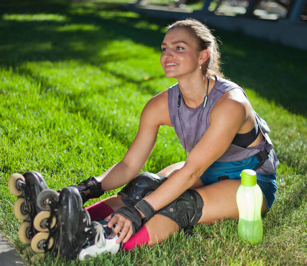 Nuori nainen, jolla on suojavarusteet ja rullaluistimet, istuu puistossa nurmikolla. Ulkoilun käsite. Harrastus. Urheiluelämäntapa - Valokuva, kuva