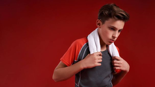 Keep the Dream Alive. A teenage boy is engaged in sport, he is looking aside while holding white towel. Isolated on red background. Fitness, training, active lifestyle concept. Horizontal shot. - Photo, image