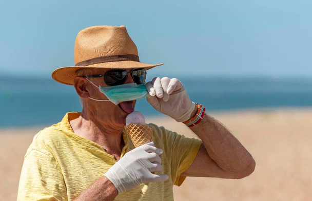 Саутсі, Портсмут, Південна Англія, Велика Британія. Травень 2020. Людина їсть морозиво на масці і гумові захисні рукавички під час спалаху Corvid-19. На березі в Саутсі.. - Фото, зображення