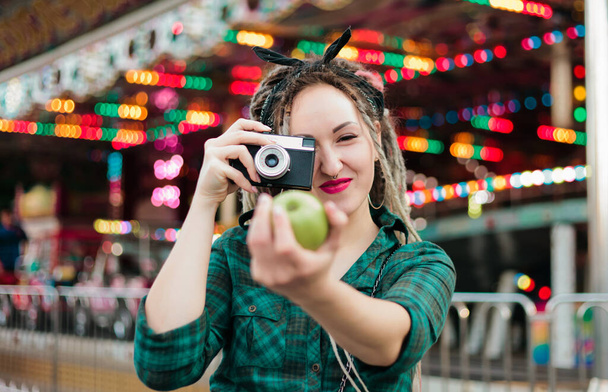 Eine ungezwungene junge Frau mit Dreadlocks fotografiert mit Retro-Kamera im Freizeitpark. - Foto, Bild