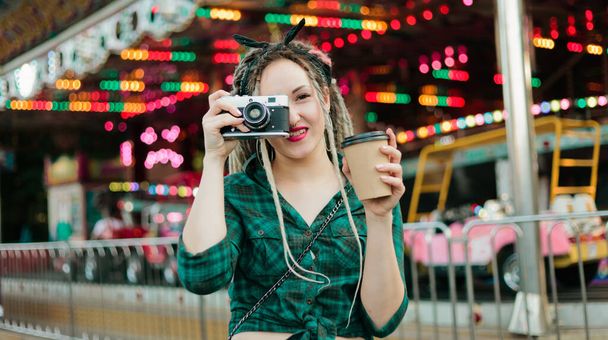 Egy nem hivatalos fiatal nő rasztával fényképeket készít retro kamerával a vidámparkban.. - Fotó, kép