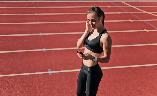 Young fit woman in sportswear listening to music with earphones and use smartphone on red-coated stadium track outdoors - Фото, зображення