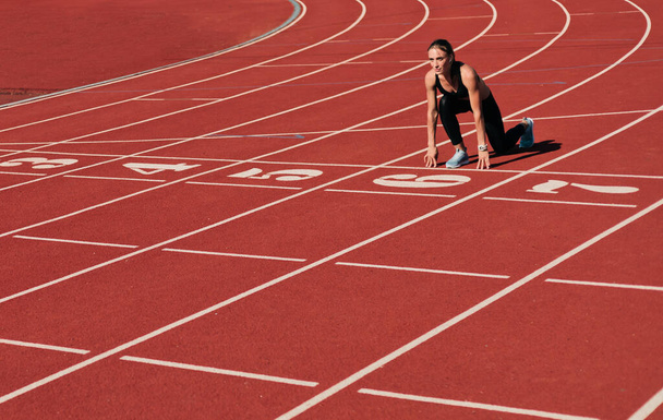 Νεαρή δρομέας γυναίκα στο αθλητικό ντύσιμο ετοιμάζεται να τρέξει σπριντ σε χαμηλή εκκίνηση στο γήπεδο κομμάτι με κόκκινο με επικάλυψη σε φωτεινή ηλιόλουστη μέρα - Φωτογραφία, εικόνα
