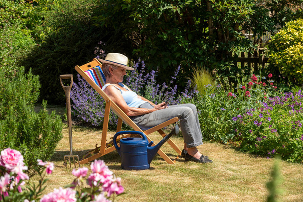 Hampshire, Anglia, Egyesült Királyság. 2020. Egy nő, aki szünetelteti a kertészkedést azzal, hogy egy nyugágyon ül egy forró nyári napon. - Fotó, kép