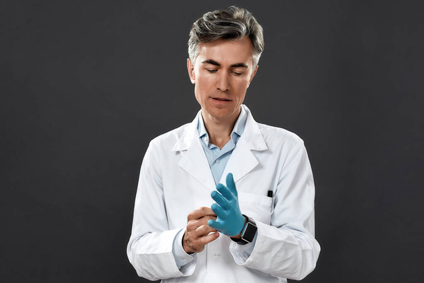 Portrait of a mature male doctor in medical uniform putting on protective sterile blue gloves while standing against black background - Foto, immagini