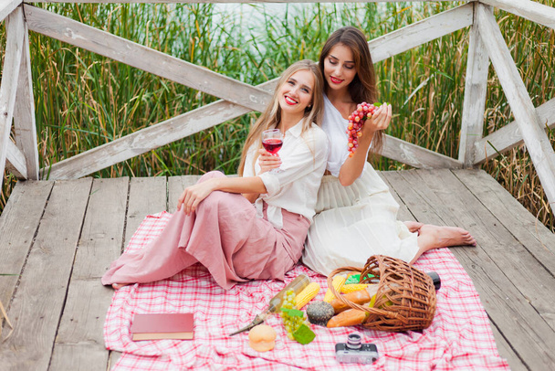 Dwie wesołe młode kobiety urządzają piknik na świeżym powietrzu w letni dzień. Dwie dziewczyny w stylu retro vintage ubrania spędzają czas razem na stawie molo. - Zdjęcie, obraz