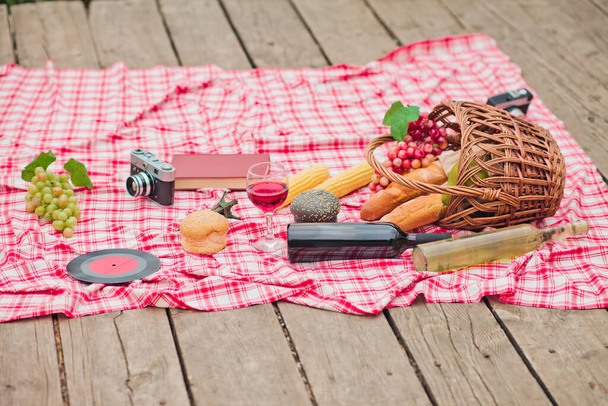 French retro style picnic. Basket with fruit, baguette, book, retro camera on a checkered red tablecloth outdoors - Fotó, kép