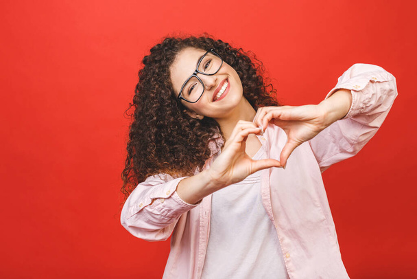 Portrét usmívající se mladé kudrnaté ženy ukazující gesto srdce se dvěma rukama a dívající se na fotoaparát izolované přes červené pozadí. - Fotografie, Obrázek