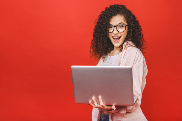 Portrait of an excited curly young girl holding laptop computer isolated over red background. - 写真・画像