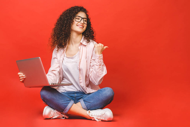 Glückliche junge lockige schöne Frau sitzt auf dem Boden mit überkreuzten Beinen und mit Laptop auf rotem Hintergrund. - Foto, Bild