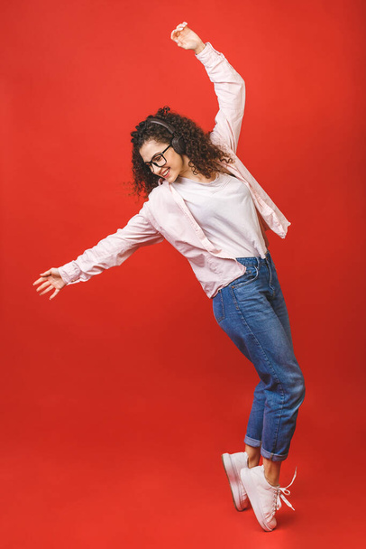 Full length portrait of a crazy cheerful girl in casual in earphones listening to music while dancing isolated over red background.  - Photo, image