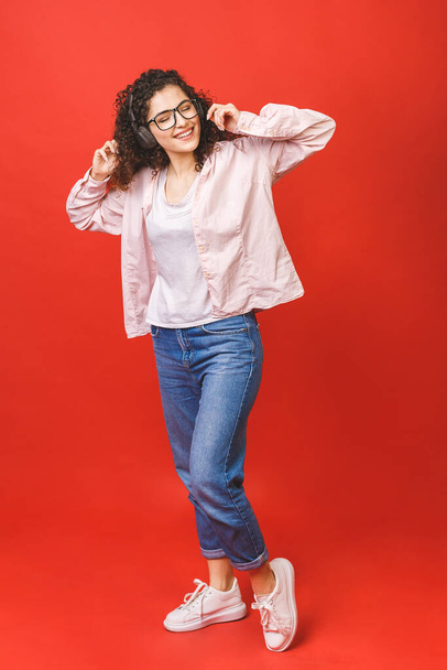 Full length portrait of a crazy cheerful girl in casual in earphones listening to music while dancing isolated over red background.  - Foto, afbeelding