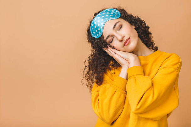 Waist up portrait of cheerful young beautiful curly smiling girl in sleeping mask. Isolated on beige background. - Фото, изображение