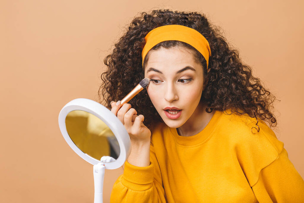Perfect view. Morning routine. Joyful young curly woman is sapplying foundation cream on her skin. She is looking at mirror in her hand. Isolated on beige background - 写真・画像