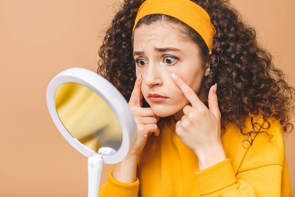 Beauty portrait of an astonished shocked amazed attractive curly woman examining her face while holding a mirror isolated over beige background. - Fotoğraf, Görsel