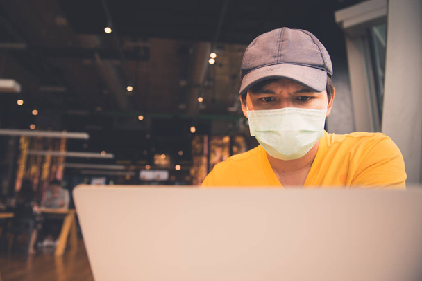 Un hombre con camisa amarilla y máscara médica usando portátil y teléfono inteligente mientras está sentado en coffeeshop
 - Foto, Imagen