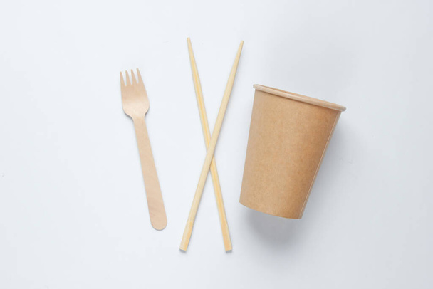 Ympäristöystävälliset ruokailuvälineet. Kiinalaisia syömäpuikkoja, puinen haarukka, käsityöpaperimuki valkoisella pohjalla. Minimalismi ekologinen konsepti. - Valokuva, kuva
