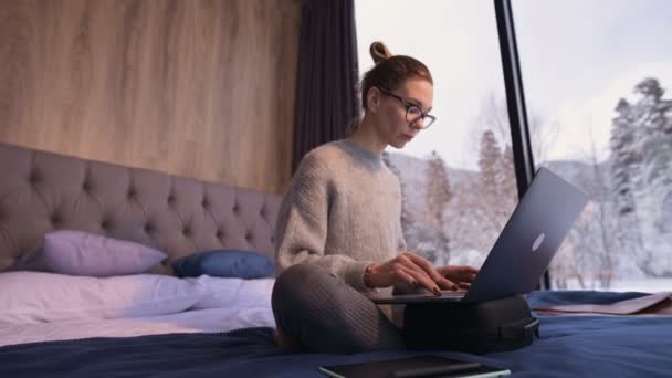 A woman freelancer with glasses sits on a bed in a country house with panoramic windows behind which there is a snowy forest. Girl with laptop computer does remotely work while isolated - Materiaali, video