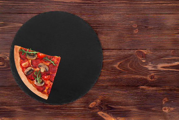Un cuarto de pizza italiana caliente con salami, rúcula, tomates cherry, champiñones y texas mezcla de especias, en un plato redondo de pizarra que está sobre fondo de madera marrón, vista superior
 - Foto, imagen